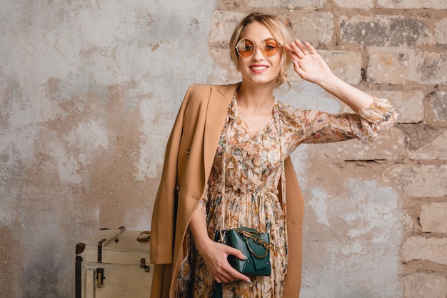 Portret van aantrekkelijke glimlachende stijlvolle blonde vrouw in beige jas wandelen in straat tegen vintage muur