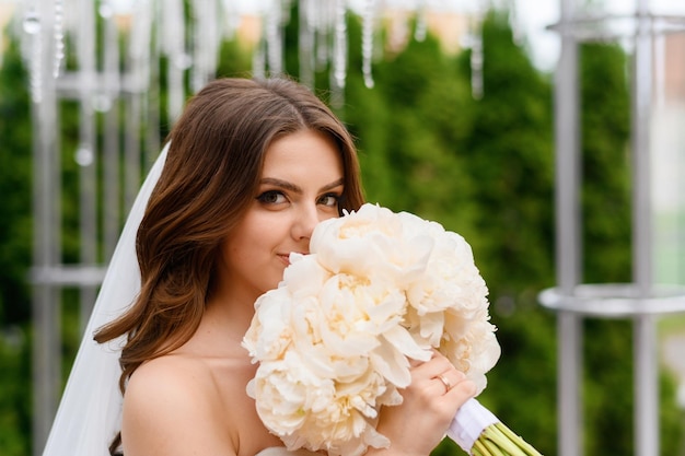 Gratis foto portret van aantrekkelijke jonge bruid vrouw in jurk met open mouwen boeket van pioenrozen houden in de buurt van mooi gezicht met naakt bruiloft make-up mooie bruid in sluier buitenshuis