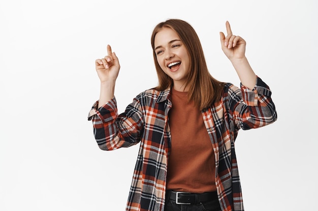 Portret van aantrekkelijke jonge vrouw dansen en lachen, glimlachen en handen opsteken, dansen op geweldige muziek, plezier hebben, staande tegen een witte achtergrond