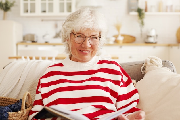 Gratis foto portret van aantrekkelijke stijlvolle europese vrouwelijke gepensioneerde m / v in ronde bril bedrijf boek, kunstgeschiedenis studeren door haarzelf, leren bij pensionering, kijken door pagina's met stralende glimlach