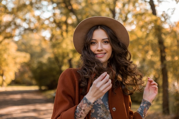 Portret van aantrekkelijke stijlvolle lachende vrouw met lang krullend haar wandelen in park gekleed in warme bruine jas herfst trendy mode, Streetstyle hoed dragen