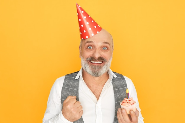 Portret van aantrekkelijke succesvolle senior bebaarde zakenman dragen kegel hoed uiting van opwinding, genieten van verjaardagsfeestje, cupcake met een kaars te houden