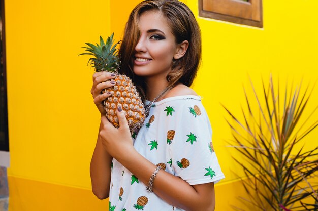 Portret van aantrekkelijke vrouw in gepint t-shirt op zomervakantie met ananas, fruit dieet detox, gebruinde huid, helder gele achtergrond