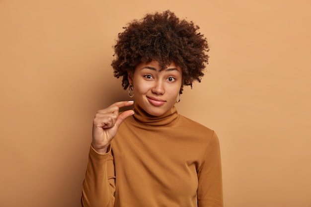 Portret van aantrekkelijke vrouw met krullend afrohaar, vormt klein en klein ding, praat over grootte, gekleed in casual bruine trui