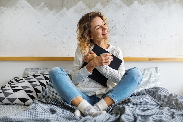 Portret van aantrekkelijke vrouw met krullend blond haar zittend op bed, boek en koffie in de beker te houden