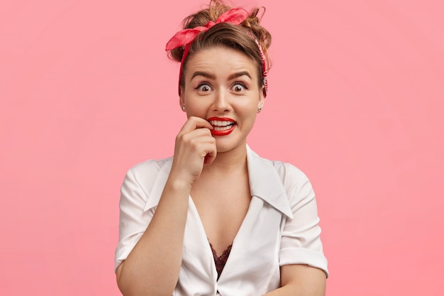 Portret van aantrekkelijke vrouw met make-up, houdt de vinger bij de mond, heeft witte, gelijkmatige tanden, draagt een hoofdband, witte blouse, geïsoleerd over roze