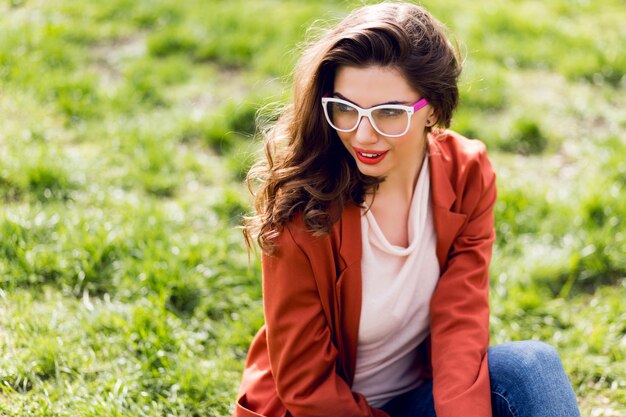 Portret van aantrekkelijke vrouw met volle lippen, brillen, rode jas, golvend kapsel zittend op groen gras in zonnige lente park en glimlachen