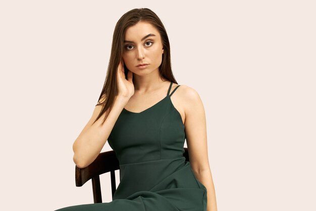 Portret van aantrekkelijke zelfverzekerde jonge blanke vrouw, gekleed in groene riem jurk poseren op stoel, aanpassen van haar lange bruine haar, serieus kijken