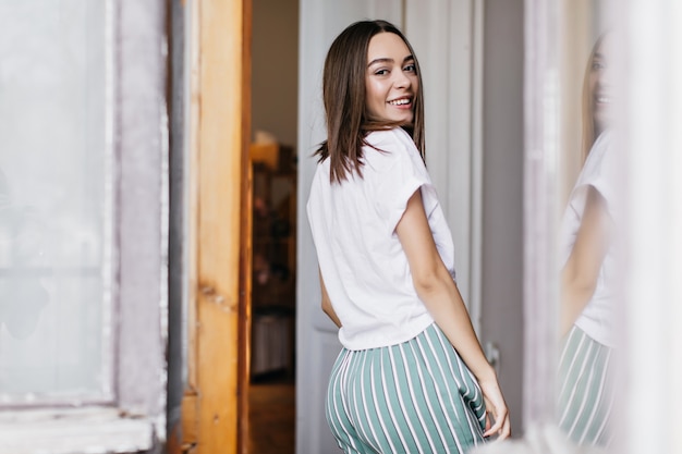 Portret van achterkant lachende vrouw in wit t-shirt plezier thuis. blij bruinharige meisje ontspannen in de ochtend.