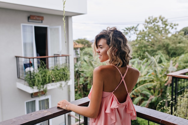 Portret van achterkant van prachtige gelooide vrouw in roze kledij kijken naar bomen. Buiten foto van romantische blonde meisje permanent op balkon en genieten van uitzicht op de stad.