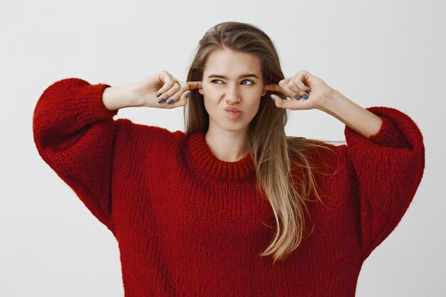 Portret van afgeleid ongemakkelijk charmant Kaukasisch meisje in losse trui, oren met wijsvingers bedekkend, naar links kijkend en grijnzend van afkeer, luid vreselijk geluid hoorend