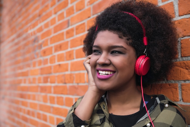 Portret van Afro-Amerikaanse Latijns-vrouw glimlachend en luisteren naar muziek met koptelefoon tegen bakstenen muur. Buitenshuis.