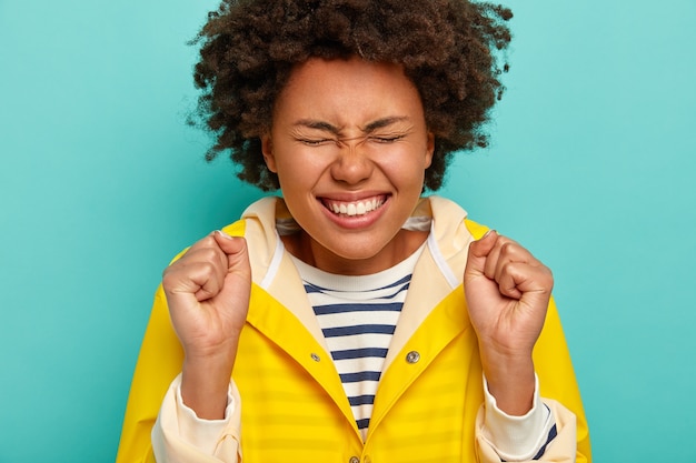 Gratis foto portret van afro-meisje viert iets, glimlacht breed, toont witte tanden, gekleed in gestreepte trui en gele regenjas, roept vrolijk, geïsoleerd op blauwe achtergrond