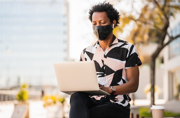 Portret van afro toeristische man met behulp van zijn laptop en het dragen van een beschermend masker terwijl hij buiten zit