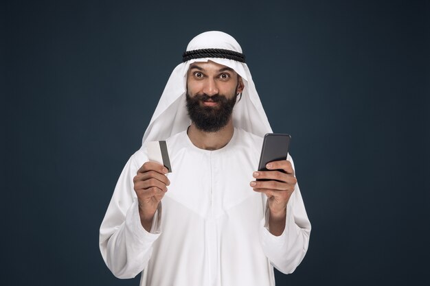 Portret van Arabische Saoedische zakenman op donkerblauwe studioachtergrond. Man met smartphone voor het betalen van rekening, online winkelen of wedden. Concept van zaken, financiën, gezichtsuitdrukking, menselijke emoties.