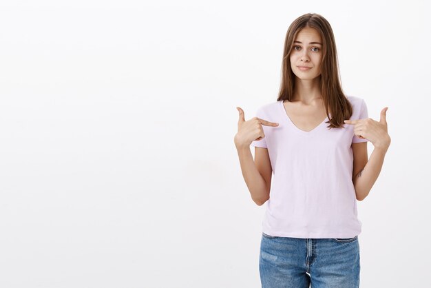 Portret van arrogante en zelfverzekerde aantrekkelijke brunette die cool handelt terwijl ze naar zichzelf wijst, lippen tuitend, ontevreden is iemand uit de lagere klasse vroeg haar nummer over een witte muur