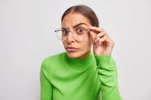 Gratis foto portret van attente jonge europese vrouw houdt hand op rand van bril heeft gerichte blik op camera luistert iets met serieuze uitdrukking draagt groene trui geïsoleerd op witte achtergrond