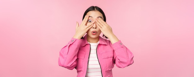 Portret van Aziatisch meisje gluurt met opwinding door vingers bedekt ogen die verrassing zien die over roze achtergrond staat