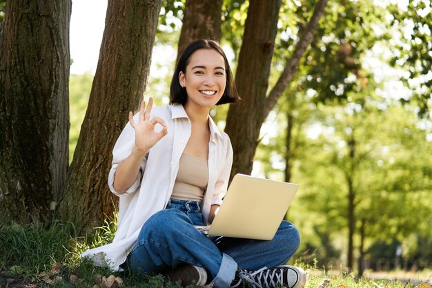 Portret van aziatische jonge studente die huiswerk doet die in parkzitting naast boom met laptop werken