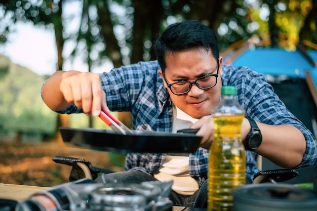 Portret van aziatische reiziger man bril varkensvlees steak frituren bbq in braadpan pan of pot op een camping buiten koken reizen camping lifestyle concept