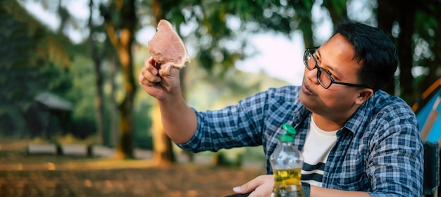 Portret van Aziatische reiziger man bril varkensvlees steak frituren BBQ in braadpan pan of pot op een camping buiten koken reizen camping lifestyle concept