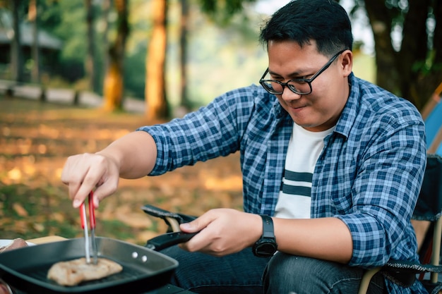 Portret van Aziatische reiziger man bril varkensvlees steak frituren BBQ in braadpan pan of pot op een camping buiten koken reizen camping lifestyle concept