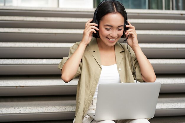 Portret van Aziatische vrouw digitale nomade meisje met behulp van laptop en luistert naar muziek buitenshuis Jonge student werkt op de computer en glimlacht