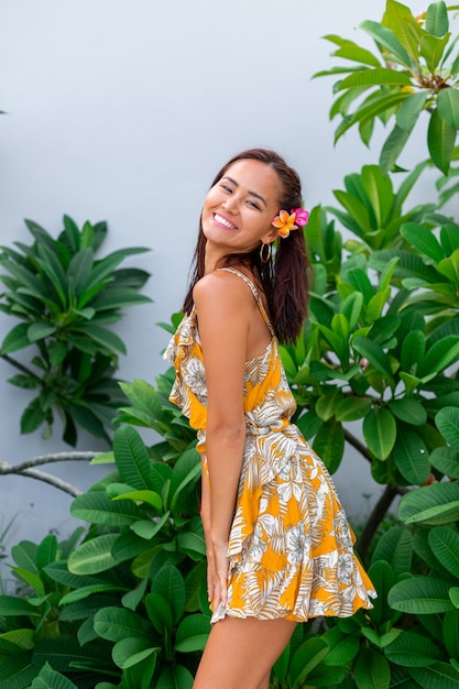 Portret van Aziatische vrouw in gele zomerjurk staat met plumeria Thaise bloem in haar en ronde oorbellen