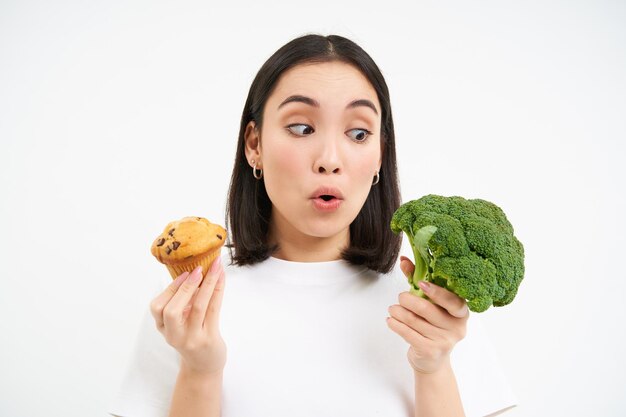 Portret van aziatische vrouw kijkt opgewonden naar broccoli met cupcake witte achtergrond