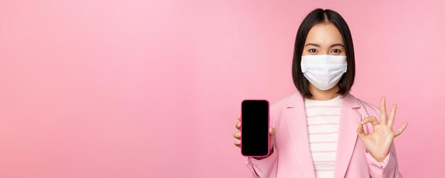 Portret van aziatische zakenvrouw met medisch gezichtsmasker covid19 met smartphonescherm en oke teken roze achtergrond