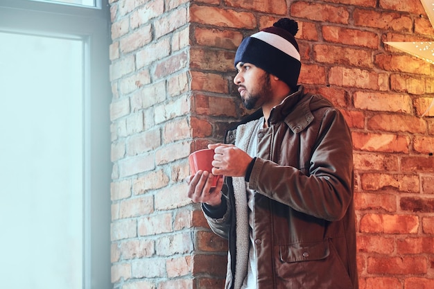 Portret van bebaarde indiase hipster man gekleed in een warme jas en een hoed, drinkt koffie uit een rode kop in de buurt van de muur van een rode baksteen.