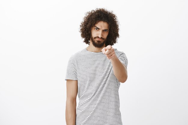 Portret van beledigde verontwaardigde knappe Spaanse man met baard en afro-kapsel, wijzend met de schuld naar voren, fronsend en ontevreden onder het voorhoofd kijkend