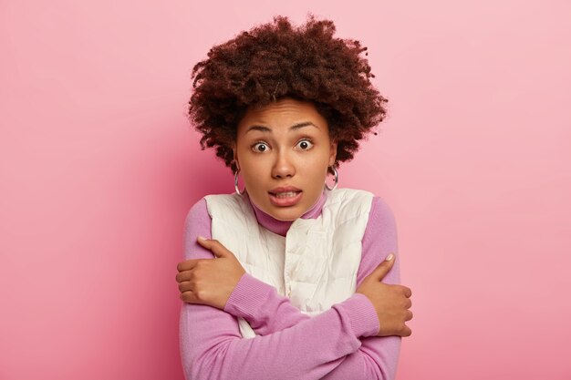 Portret van bevende gekrulde vrouw rilt van de kou, kruist armen, klemt tanden, kijkt verrassend naar de camera, geïsoleerd op roze achtergrond