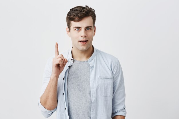 Portret van blanke man met grote ogen en donker haar dat door iets wordt geschokt, gekleed in een blauw shirt boven een t-shirt, wijzend met zijn vinger naar de blinde muur met kopie ruimte voor uw advertentie-inhoud