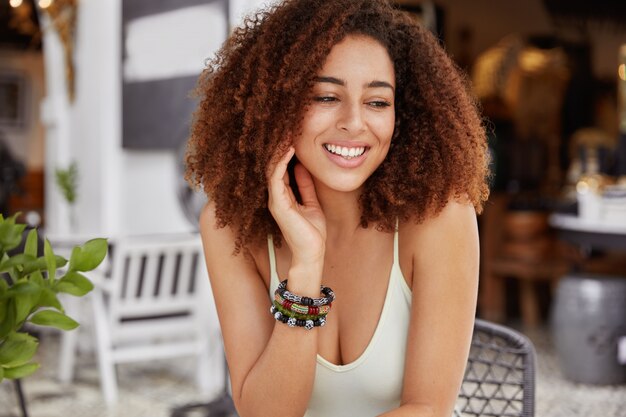 Portret van blij Afro-Amerikaanse vrouw heeft een positieve glimlach, heeft borstelige haarstijl kijkt met dromerige uitdrukking weg zit tegen cafetaria interieur.