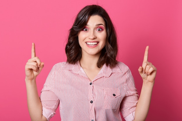 Portret van blij meisje dat gestreept overhemd draagt, direct camera bekijkt, wijzend beide wijsvingers omhoog, jonge vrouw met groot idee, die zich tegen roze muur bevindt.