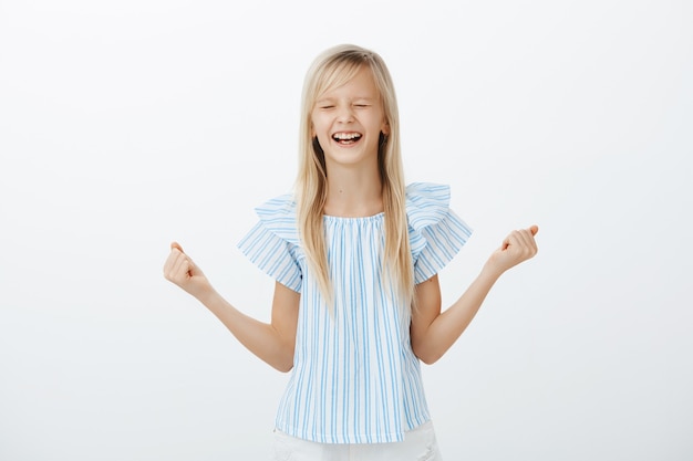 Portret van blij opgewonden mooi meisje met blond haar in blauwe blouse, gebalde vuist, ogen sluiten en schreeuwen van gejuich en geluk, vreugdevol zijn, eerste plaats winnen op evenement