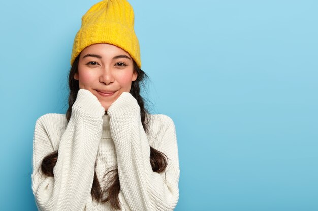 Portret van blij schattige Aziatische vrouw houdt handen onder de kin, gekleed in oversized witte trui en gele hoed, geniet van comfort en rustige huiselijke sfeer