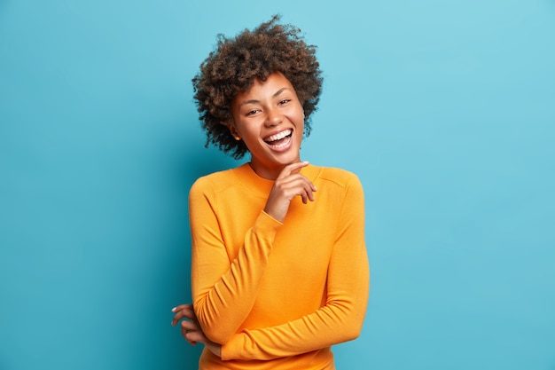 Portret van blije jonge vrouw lacht vrolijk houdt hand op kin drukt positieve emoties uit glimlacht in het algemeen heeft zorgeloze uitdrukking draagt oranje trui geïsoleerd over blauwe muur
