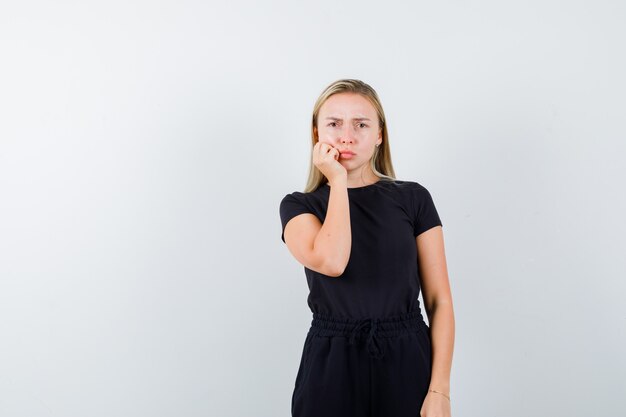 Portret van blonde dame die hand op wang in zwarte kleding houdt en somber vooraanzicht kijkt