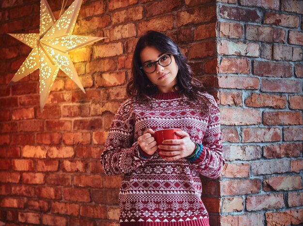 Portret van brunette vrouw gekleed in een rode trui drinkt koffie over de muur van een baksteen.