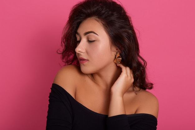 Portret van brunette vrouw met golvend haar poseren met de handen in de nek en opzij kijken, het dragen van een zwart shirt met blote schouders, geïsoleerd over roze muur.