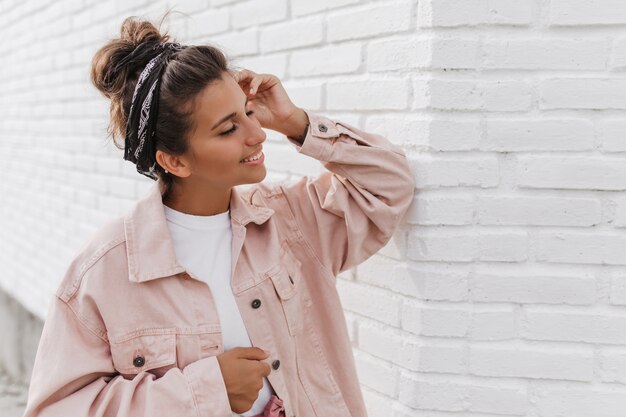 Portret van charmante donkerharige vrouw in roze jasje leunde op lichte bakstenen muur