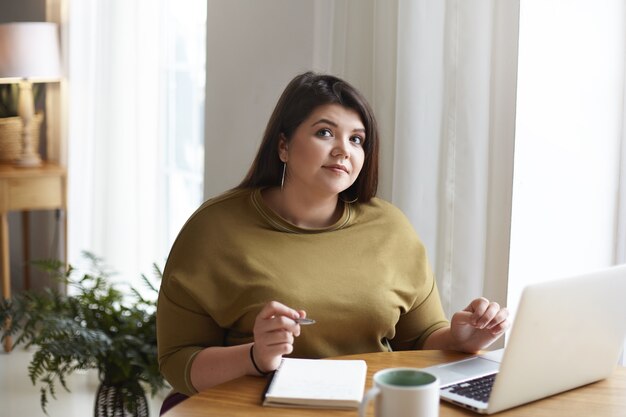 Portret van charmante elegante jonge vrouw schrijver met mollige wangen en donker haar notities maken in haar beurt met behulp van draadloze internetverbinding op generieke draagbare computer thuis kantoor