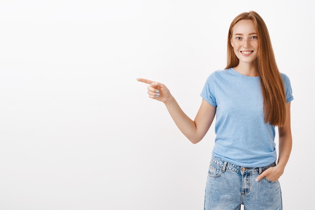 Portret van charmante roodharige jonge vrouw met sproeten hand in hand in de zak en naar links wijzend met blije beleefde glimlach