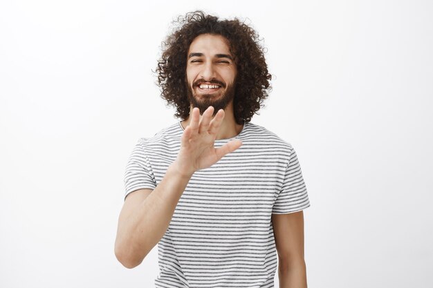 Portret van charmante zorgeloze spaanse vriend met krullend haar en baard, wuivende palm in nee, bedankt of stop gebaar en breed glimlachend met vriendelijke houding