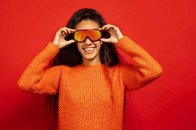 Portret van de afro-amerikaanse jonge brunette vrouw in skimasker op rode studio achtergrond. concept van menselijke emoties, gezichtsuitdrukking, verkoop, advertentie, wintersport en vakanties. glimlachen, bril dragen.