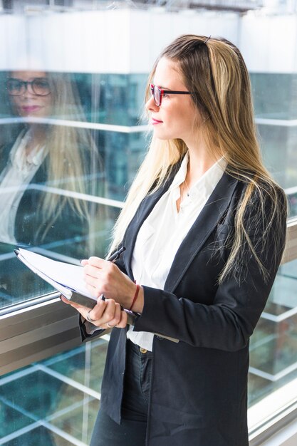 Portret van de holdingsdocument van de blonde jong vrouw die zich dichtbij het vensterglas bevinden