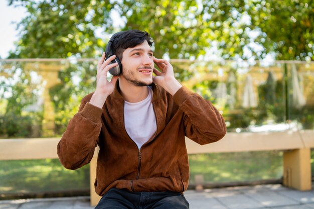 Portret van de jonge knappe man, luisteren naar muziek met een koptelefoon zittend buiten. Stedelijk concept.