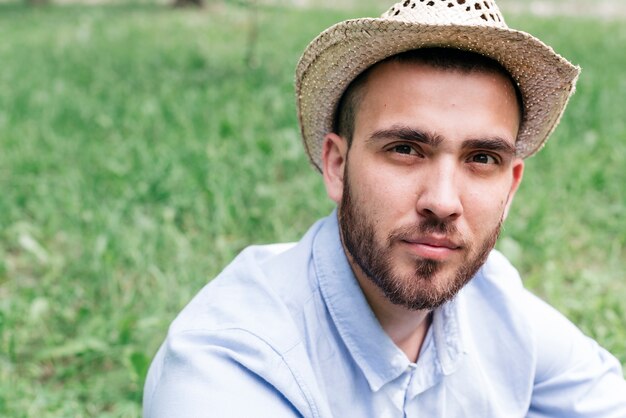 Portret van de jonge mens die hoed draagt die camerazitting op gras bekijkt